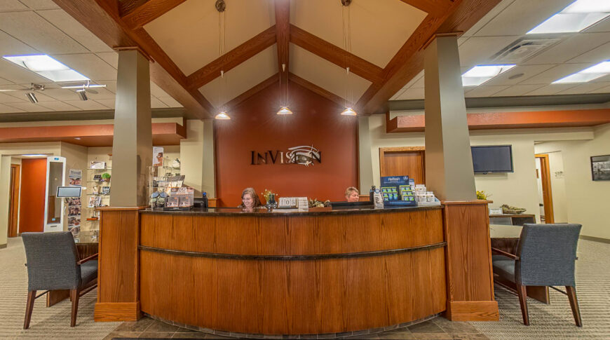 Entryway view of InVision Eye Clinic leading to curved wooden reception desk with two columns on either side holding up an A-frame arched ceiling above. Accent wall in burn orange in the background with the InVision logo.