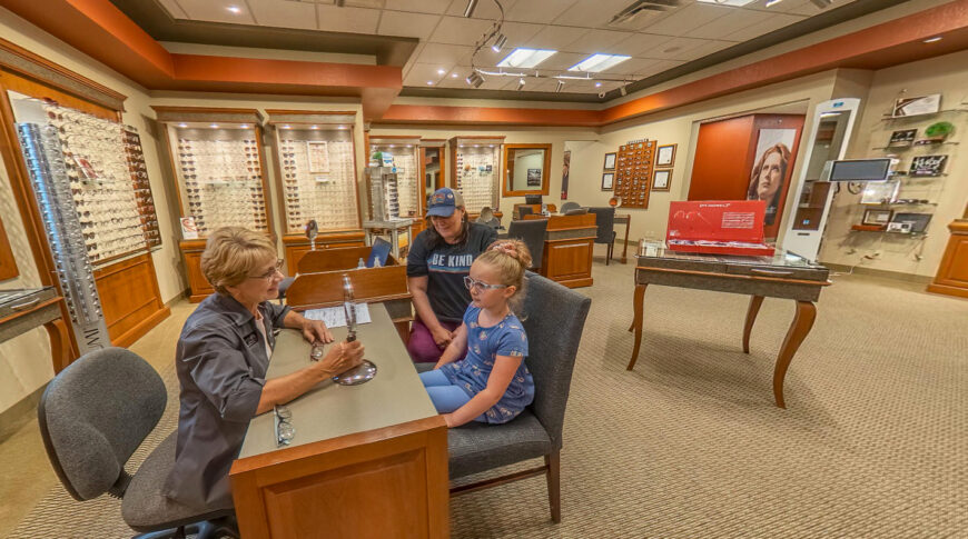 Eye glasses store with cases filled with glasses frames in the background and tables to ensure proper fit in foreground. Child and mother sitting across glass fitter at one table.