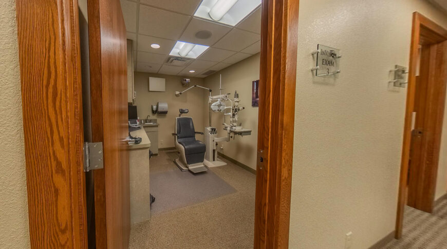 Hallway view looking in to an eye exam room with chair and examination equipment.
