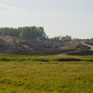 Scenic landscape of lush green wild-grown field in foreground followed by dirt field with machinery moving dirt up small hill in background.