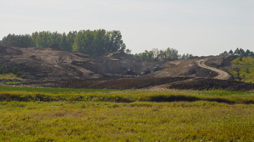 Scenic landscape of lush green wild-grown field in foreground followed by dirt field with machinery moving dirt up small hill in background.