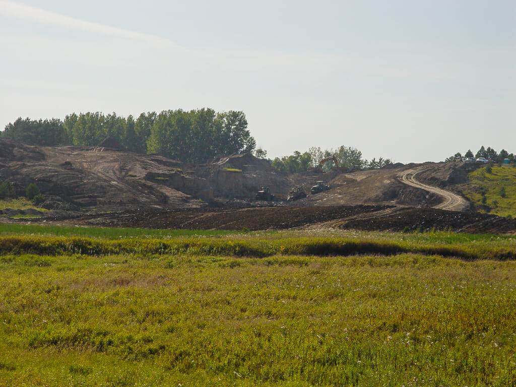 Scenic landscape of lush green wild-grown field in foreground followed by dirt field with machinery moving dirt up small hill in background.