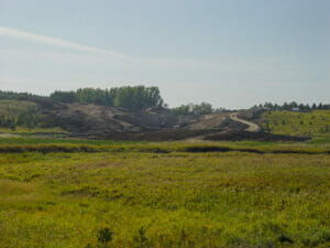 Scenic landscape of lush green wild-grown field in foreground followed by dirt field with machinery moving dirt up small hill in background.