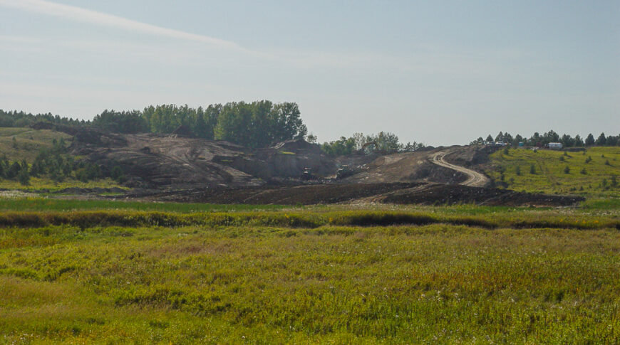 Scenic landscape of lush green wild-grown field in foreground followed by dirt field with machinery moving dirt up small hill in background.