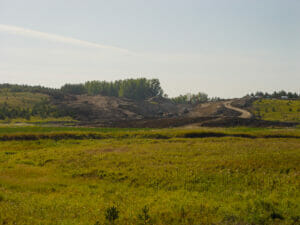Scenic landscape of lush green wild-grown field in foreground followed by dirt field with machinery moving dirt up small hill in background.