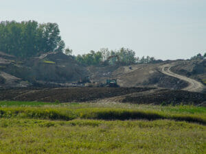 Scenic landscape of lush green wild-grown field in foreground followed by dirt field with machinery moving dirt up small hill in background.
