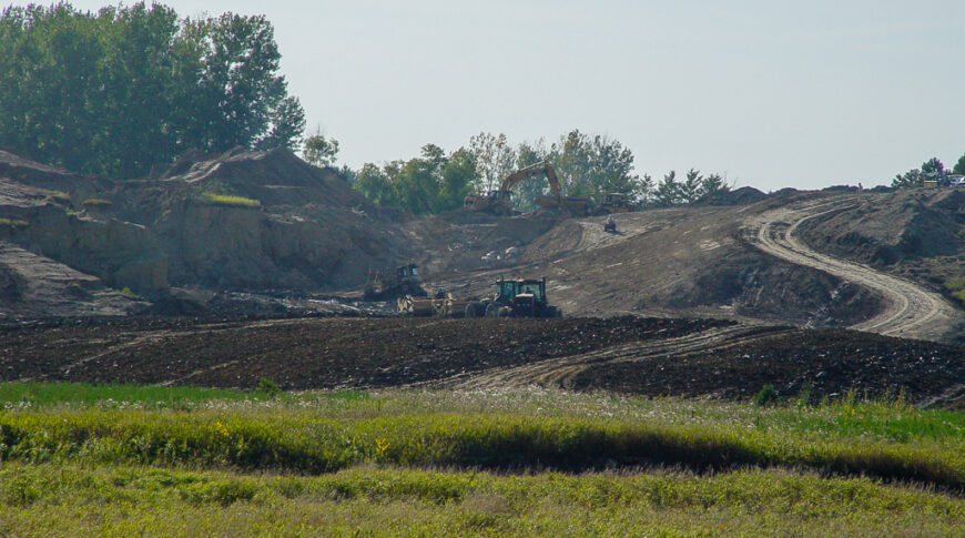 Scenic landscape of lush green wild-grown field in foreground followed by dirt field with machinery moving dirt up small hill in background.