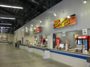 Concession windows with garbage cans spaced evenly between each window. Concrete floors beneath with black exposed beams of the grandstands above.