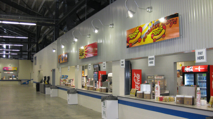 Concession windows with garbage cans spaced evenly between each window. Concrete floors beneath with black exposed beams of the grandstands above.