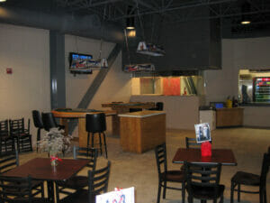 Beige cinder block room with bar to the back, gambling tables in middle and small square tables and chairs in foreground.