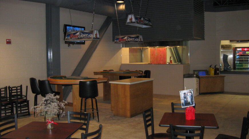 Beige cinder block room with bar to the back, gambling tables in middle and small square tables and chairs in foreground.