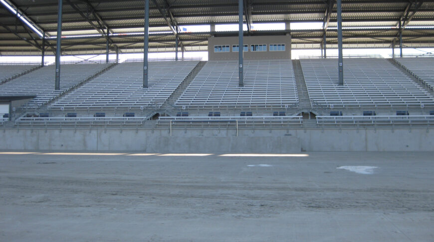 Track view of concrete ND State Fair Grandstands base with metal bleachers and steel beams upholding metal roof above.