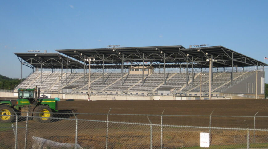 Exterior view of North Dakota State Fair Grandstands from far opposite side of the field on sunny day with blue sky in background and green tractor in foreground.