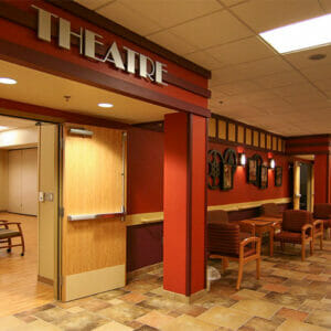 Hallway with double doors leading to room with wooden flooring and chairs throughout with an archway above and a sign that says 'Theatre."