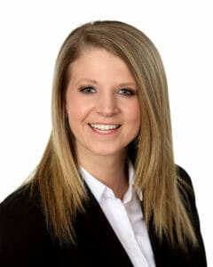 Young woman with long blonde hair smiling for professional headshot with white background wearing black jacket and white button-up shirt.
