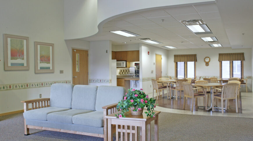 Large common room with couch seating area in foreground, small square dining tables and chairs to back right and kitchen in nook to back left.