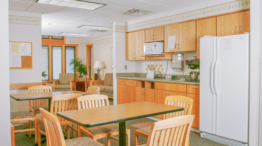 Kitchenette with cabinetry on the back right wall with two rectangular tables and chairs in foreground and living room area to background.