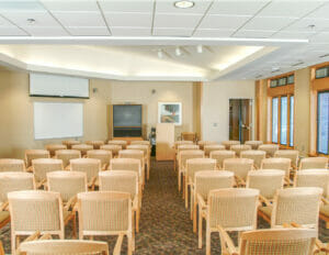 Meeting room at Project Turnabout holding about fifty wooden and fabric covered chairs with projector, white board and large tv on the back wall and wall of windows to the right wall.