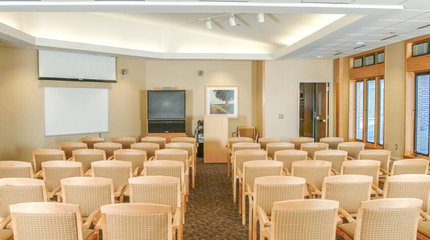 Meeting room holding about fifty wooden and fabric covered chairs with projector, white board and large tv on the back wall and wall of windows to the right wall.