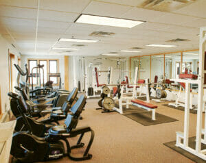 Project Turnabout gym facility room with aerobic and weight-lifting equipment throughout with paneled ceiling tiles and brown carpeting and wall of mirrors to the back.