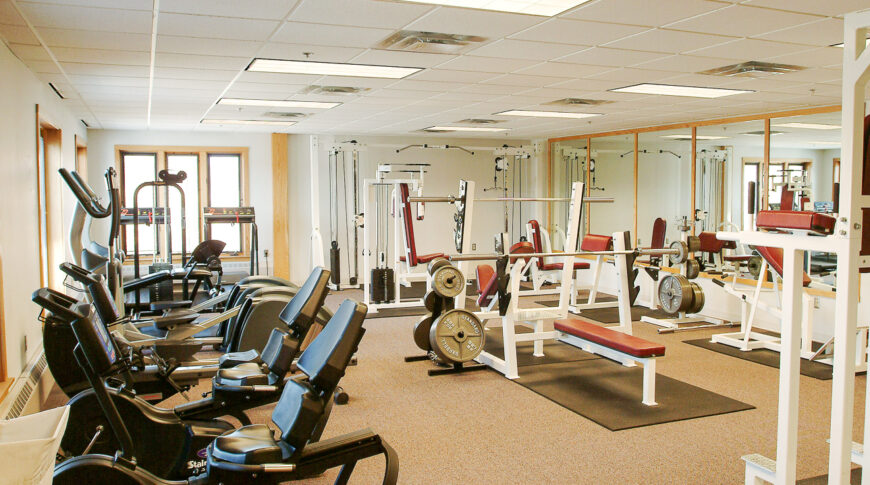Gym facility room with aerobic and weight-lifting equipment throughout with paneled ceiling tiles and brown carpeting and wall of mirrors to the back.