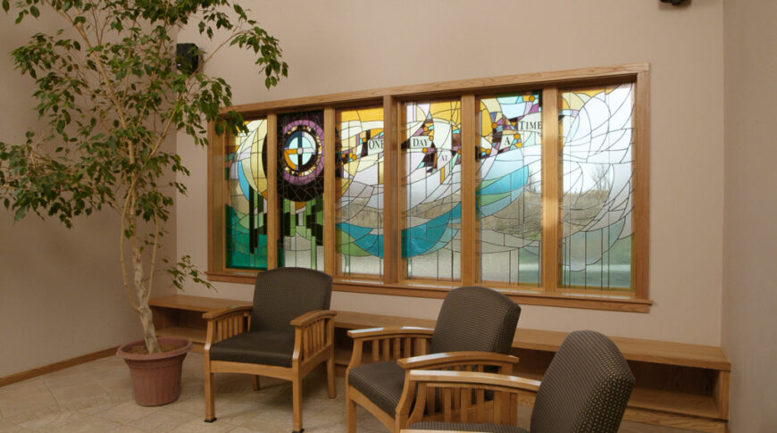 Tiled seating area with three wooden chairs with dark brown cushions, stained glass windows behind and faux birch tree in a pot to the far left.