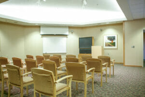 Meeting room with wooden chairs throughout with retractable projector screen over whiteboard to the back wall with TV and podium to the right of it.