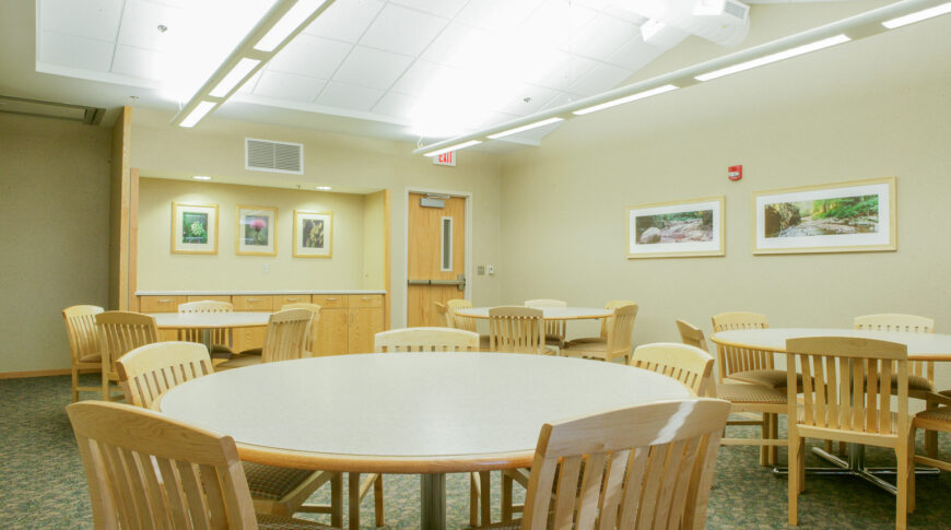 Medium-sized room with four round tables surrounded by wooden chairs with under cabinetry to far back wall and exit door to the right of said cabinetry.