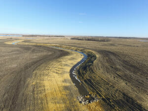 Aerial view of drainage channel snaking through brown dirt fields with rock check in foreground.
