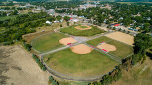 Aerial view of four baseball fields on sunny summer day with lush green trees surrounding and small city in the background.