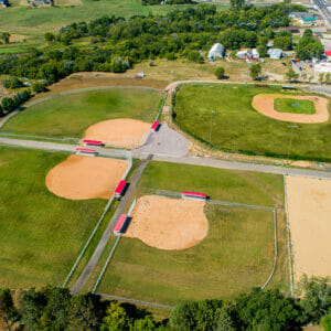Aerial view of four baseball fields on sunny summer day. Three newer fields have bright red-roofed dugouts and older field has bleacher system.