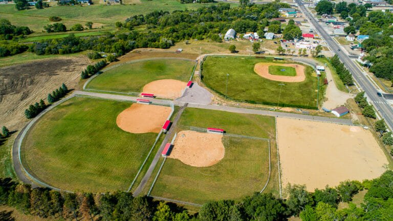 Aerial view of four baseball fields on sunny summer day. Three newer fields have bright red-roofed dugouts and older field has bleacher system.