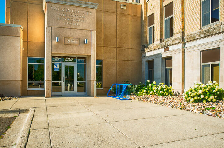 Precast concrete exterior entrance view of Wilkin County Jail building connected to the original government brick building to the right.