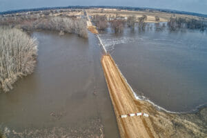 Gravel road with barricades blocking off flooded section of roadway. Overflooded river on either side of road spilling onto gravel road.