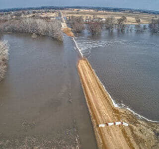 Gravel road with barricades blocking off flooded section of roadway. Overflooded river on either side of road spilling onto gravel road.