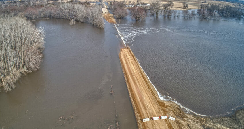 Gravel road with barricades blocking off flooded section of roadway. Overflooded river on either side of road spilling onto gravel road.