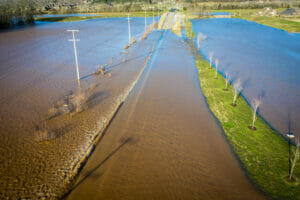 Aerial view of flooding over rural road with high water in ditches on either side and some residential housing to the background.