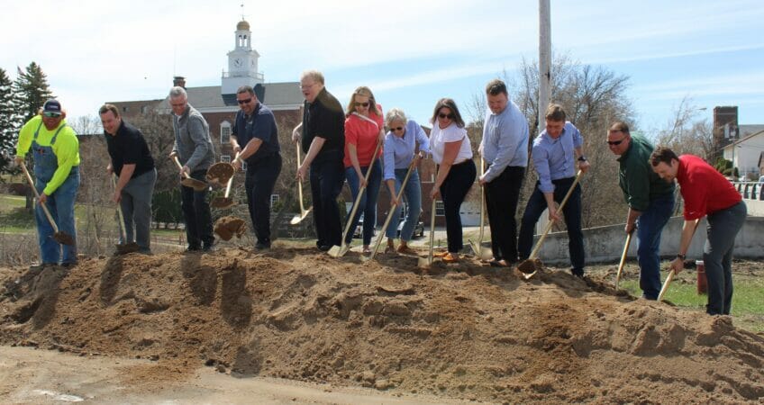 Group of people standing on sand pile shoveling scoops of sand for groundbreaking ceremony for the Fergus Falls Riverfront Phase II project.