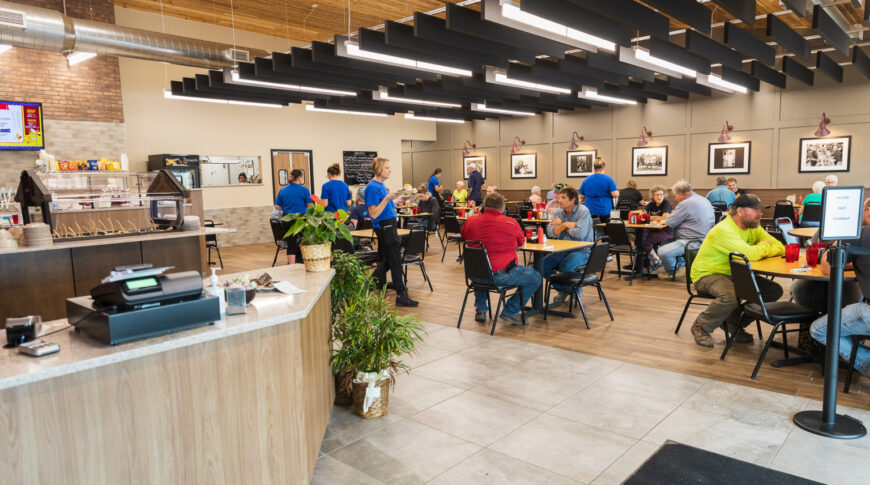 Dining area in Cenex C-Store with small square tables and black chairs with architectural accents overhead and cashier to foreground