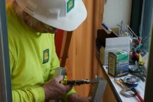 Image of old man with white hair wearing a Comstock Construction logo'd white hard hat and safety long-sleeve t-shirt cutting metal framing.
