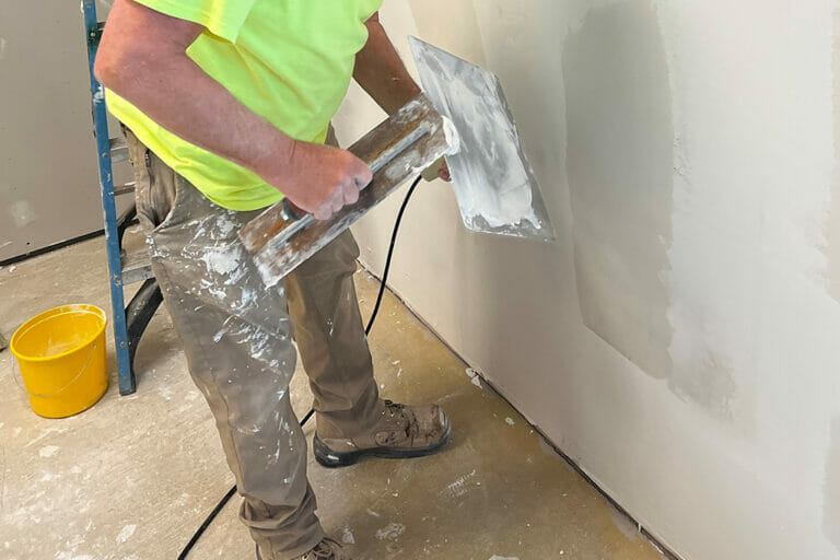 Man in yellow shirt and khaki pants mudding a drywalled wall.