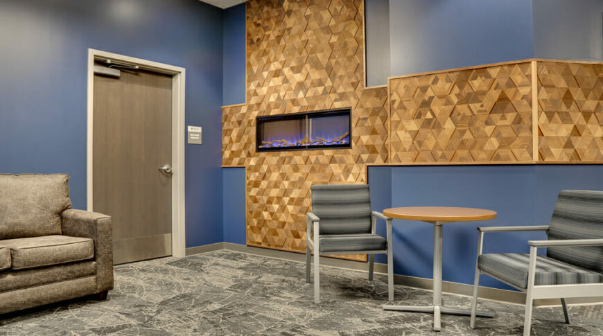 Waiting area with couch to back left wall, small table with two chairs to back right, flanked by blue accent wall with fireplace and custom cross-shaped wooden triangle accent.