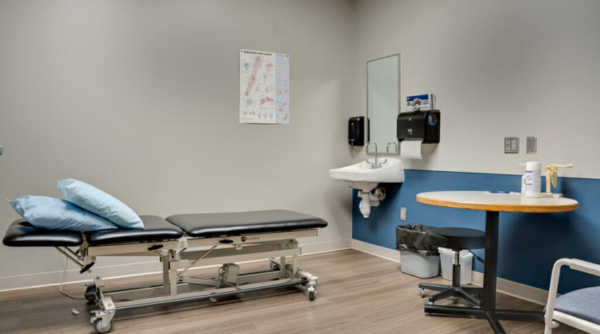 Beige exam room with exam table, seating chairs, sink and blue accent wall.