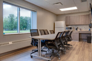 Breakroom at the LifeCare Medical Warroad with long table surrounded by 8 black chairs, window overlooking exterior grounds to the back left and kitchenette station with cabinetry, and appliances to back right.