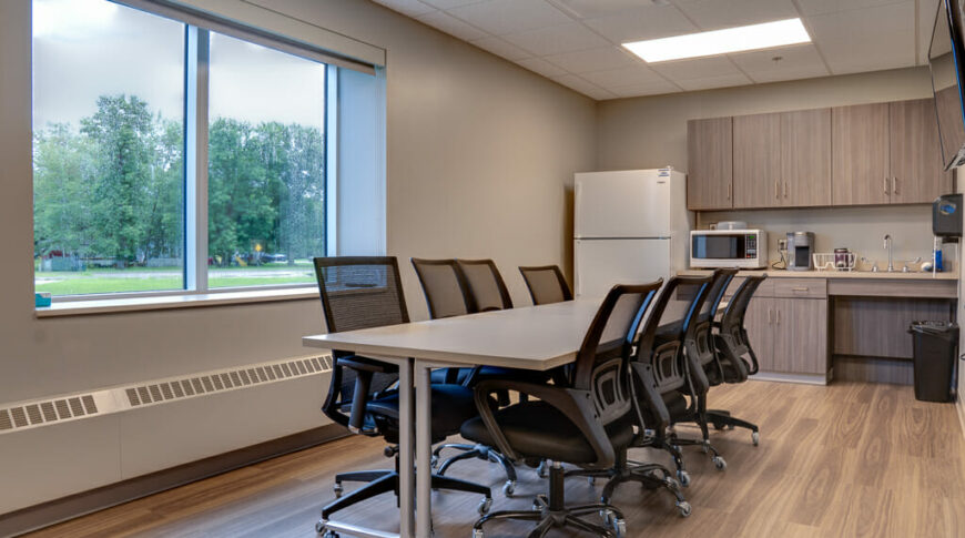 Breakroom at the LifeCare Medical Warroad with long table surrounded by 8 black chairs, window overlooking exterior grounds to the back left and kitchenette station with cabinetry, and appliances to back right.
