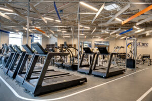 Inside fitness center with treadmills and other gym equipment in the middle surrounded by a walking track. Ceiling with exposed industrial look and beige, blue and orange acoustic panels hanging above.