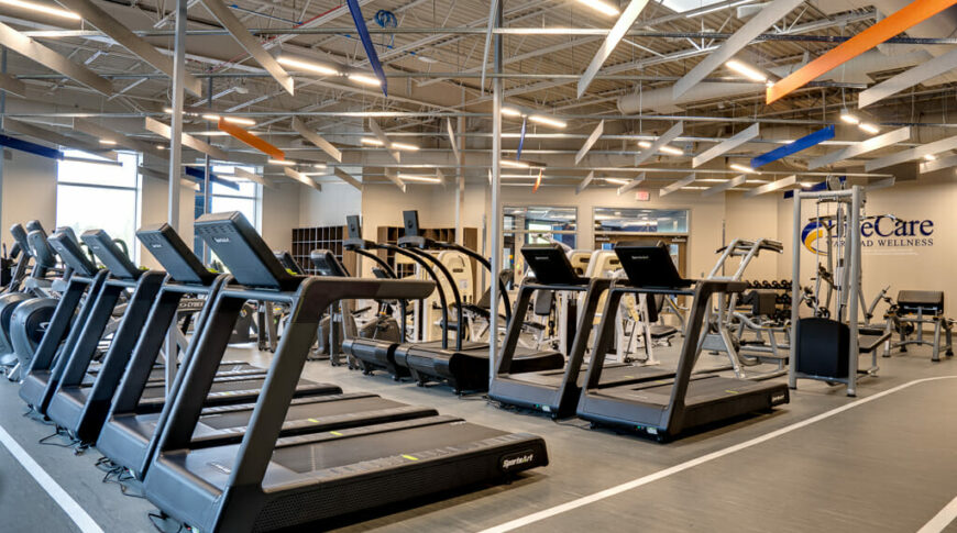 Inside fitness center with treadmills and other gym equipment in the middle surrounded by a walking track. Ceiling with exposed industrial look and beige, blue and orange acoustic panels hanging above.