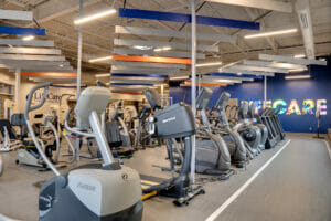 Inside of newly renovated gym with aerobic equipment in middle with vibrant blue wall to the back with lit "LIFECARE" marquee on it.