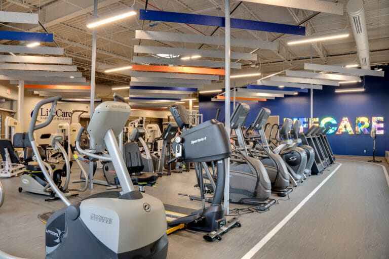 Inside of newly renovated gym with aerobic equipment in middle with vibrant blue wall to the back with lit "LIFECARE" marquee on it.
