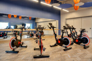 Fitness class room with chevron-designed floor, eliptical machines in foreground with back blue wall of mirrors and ballet bar. Ceiling has blue, beige, and orange hanging acoustic tiles.